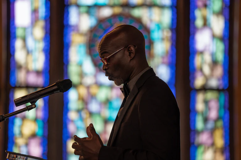A Black man in glasses and a suit looks down as he speaks into a microphone, standing in front of stained glass windows.