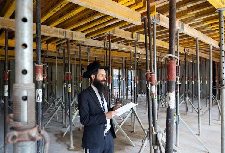A bearded man wearing a hat, suit and tie walks into a room under construction with exposed beams.