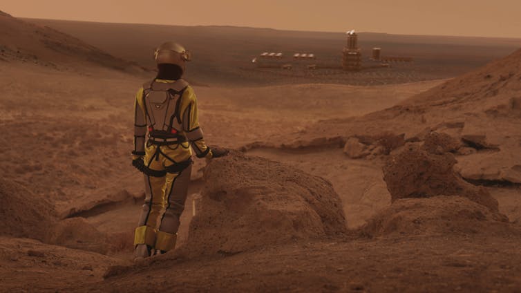 looking from behind astronaut over a ridge on a rocky landscape toward buildings