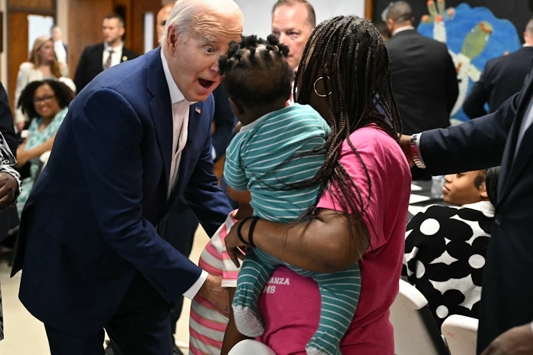 An older man with white hair and a dark suit makes a surprised face as he faces a woman with a baby.