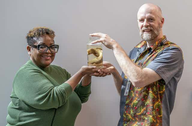 A man and woman hold up a preserved lizard in a jar