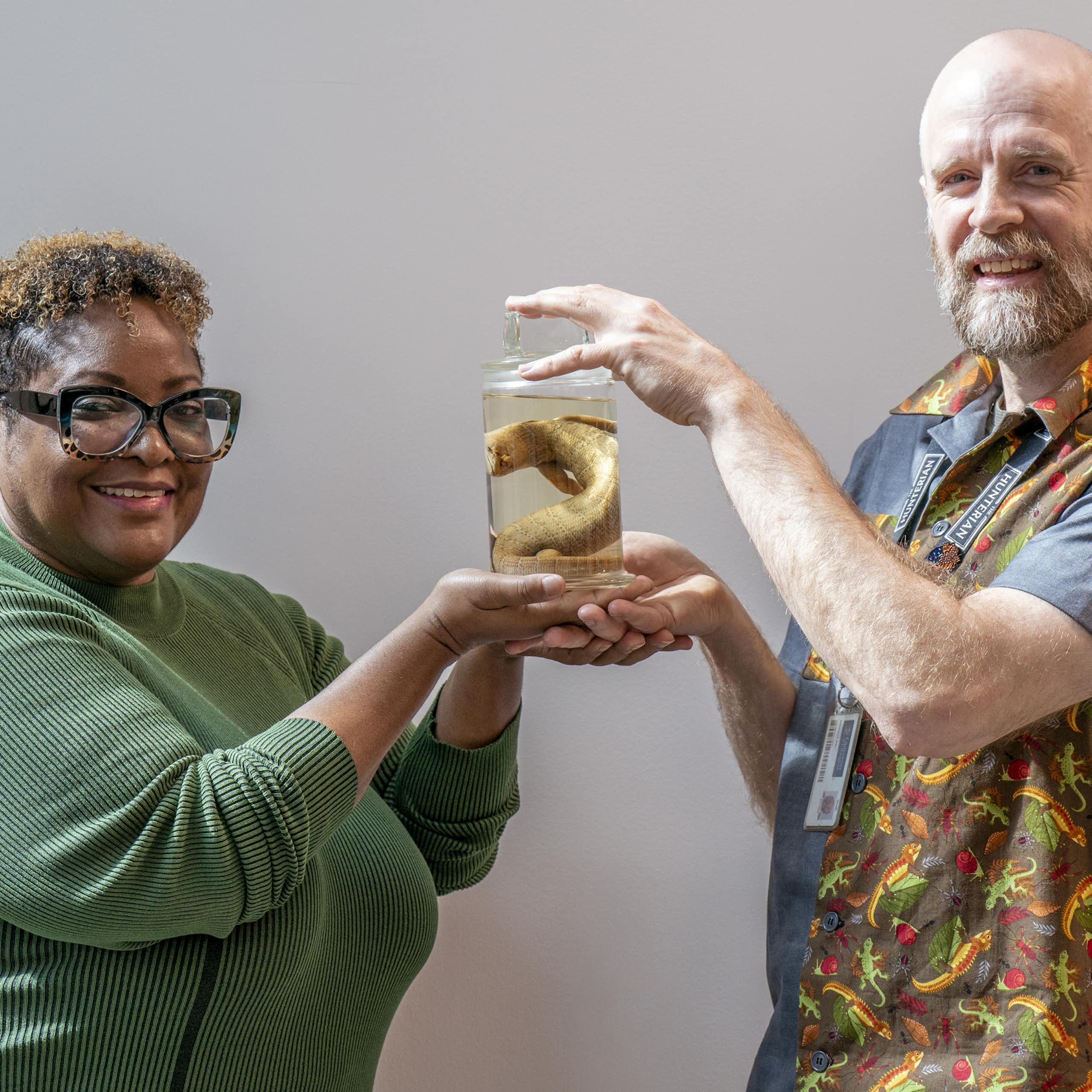 A man and woman hold up a preserved lizard in a jar