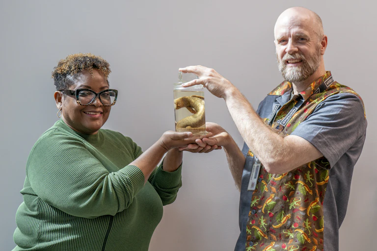 A man and woman hold up a preserved lizard in a jar