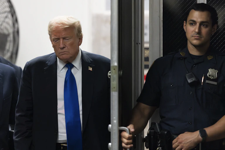 A man in a suit and tie walks through a door held by a police officer.