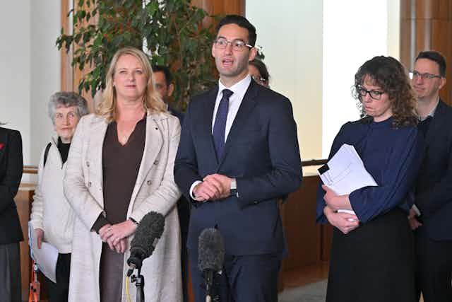 A man in a suit and a woman in a coat stand behind microphones and address the media