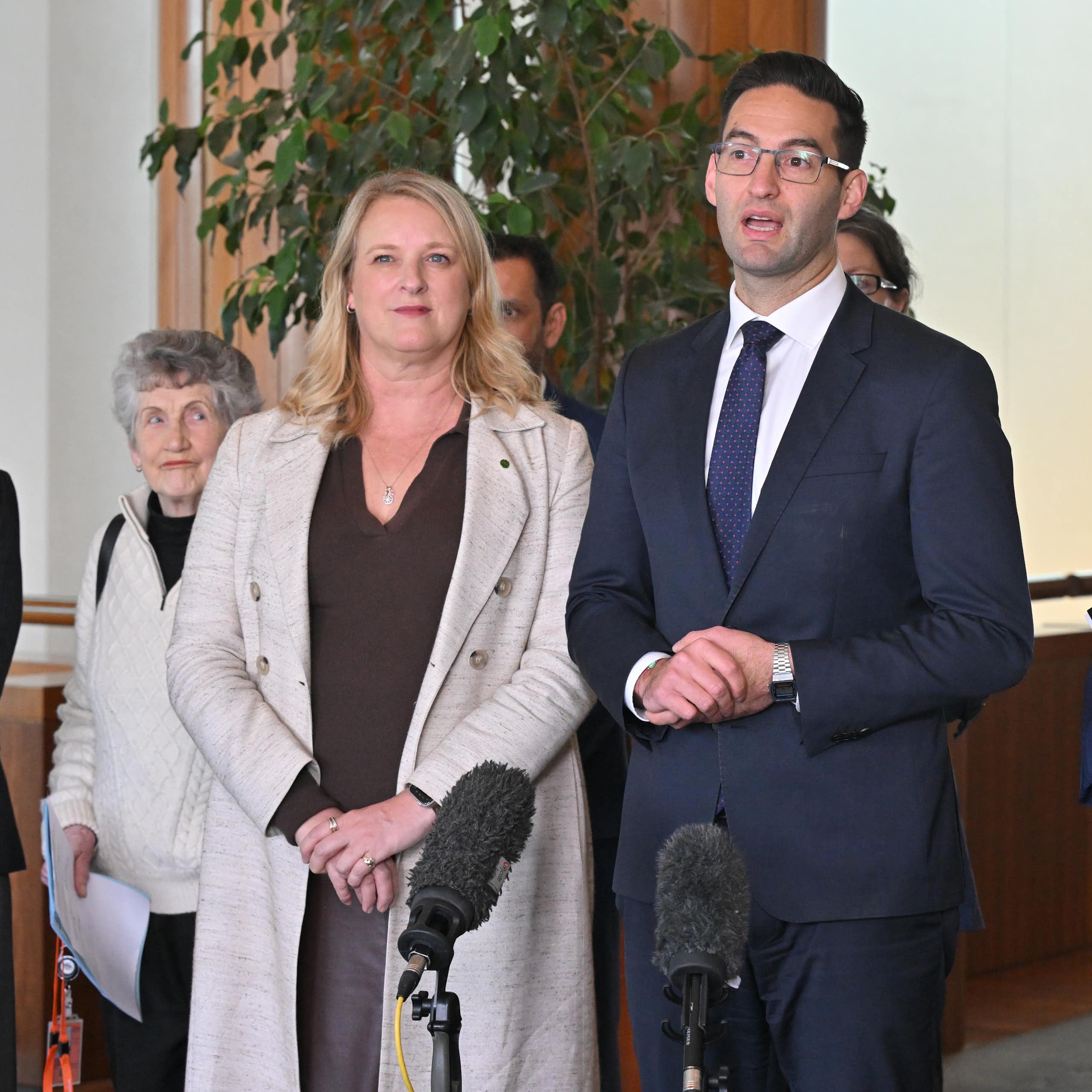 A man in a suit and a woman in a coat stand behind microphones and address the media