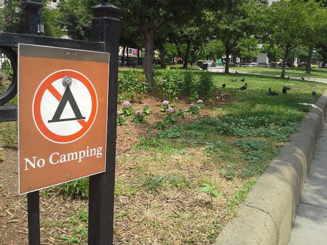 A small sign that says 'No Camping' and has a picture of a tent crossed out, positioned near a park.