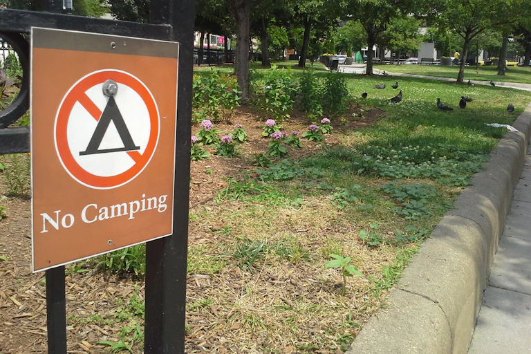 A small sign that says 'No Camping' and has a picture of a tent crossed out, positioned near a park.