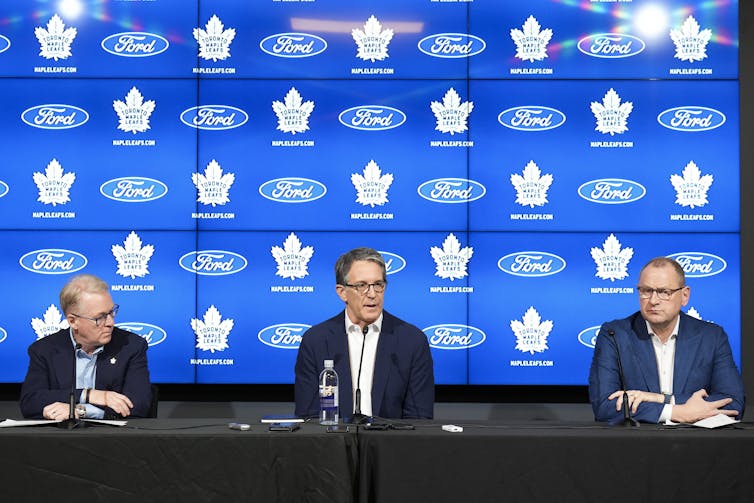 Three men in suit jackets sit at a long table in front of screens displaying the logos of the Toronto Maple Leafs and Ford Motors