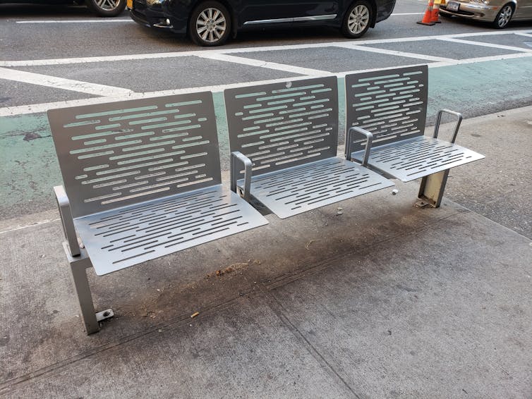A grey bench with a perforated pattern and armrests that divide it into three seats stands on a sidewalk.