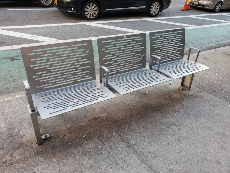 A gray bench with pinhole designs and armrests separating it into three seats, positioned on a sidewalk.