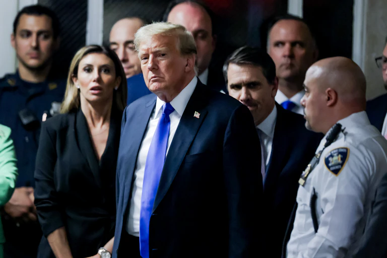 A man ina. dark blue suit with a white shirt and a white tie has a solemn face as he walks, surrounded by a security guard, a police officer and other people in formal clothing.