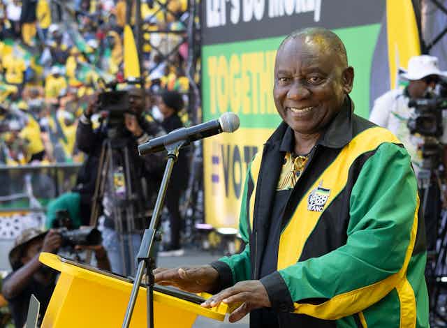 A man dressed in a green, yellow and black jacket standing in front of a microphone smiling at the camera.