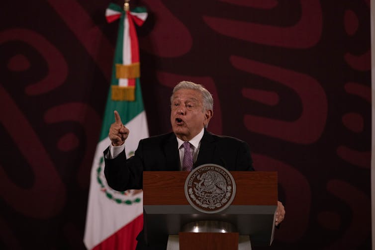 A man with white hair dressed in a suit delivering a speech in front of a Mexican flag.