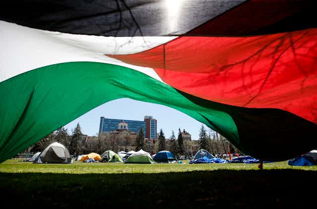 A tent encampment is pictured under a large Palestinian flag in the foreground.