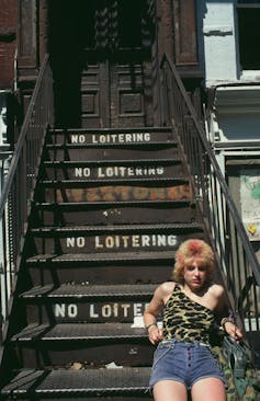 A young woman with dyed hair and a leopard print top leans back on metal steps.