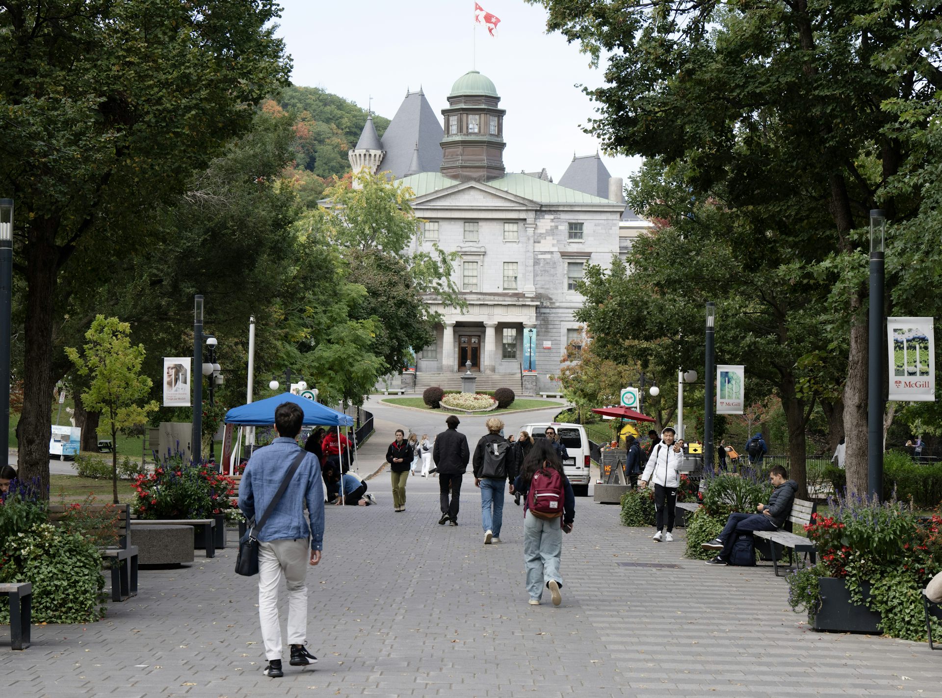 Students carrying book bags and knapsacks walk toward an ornate stone building on a treed path.