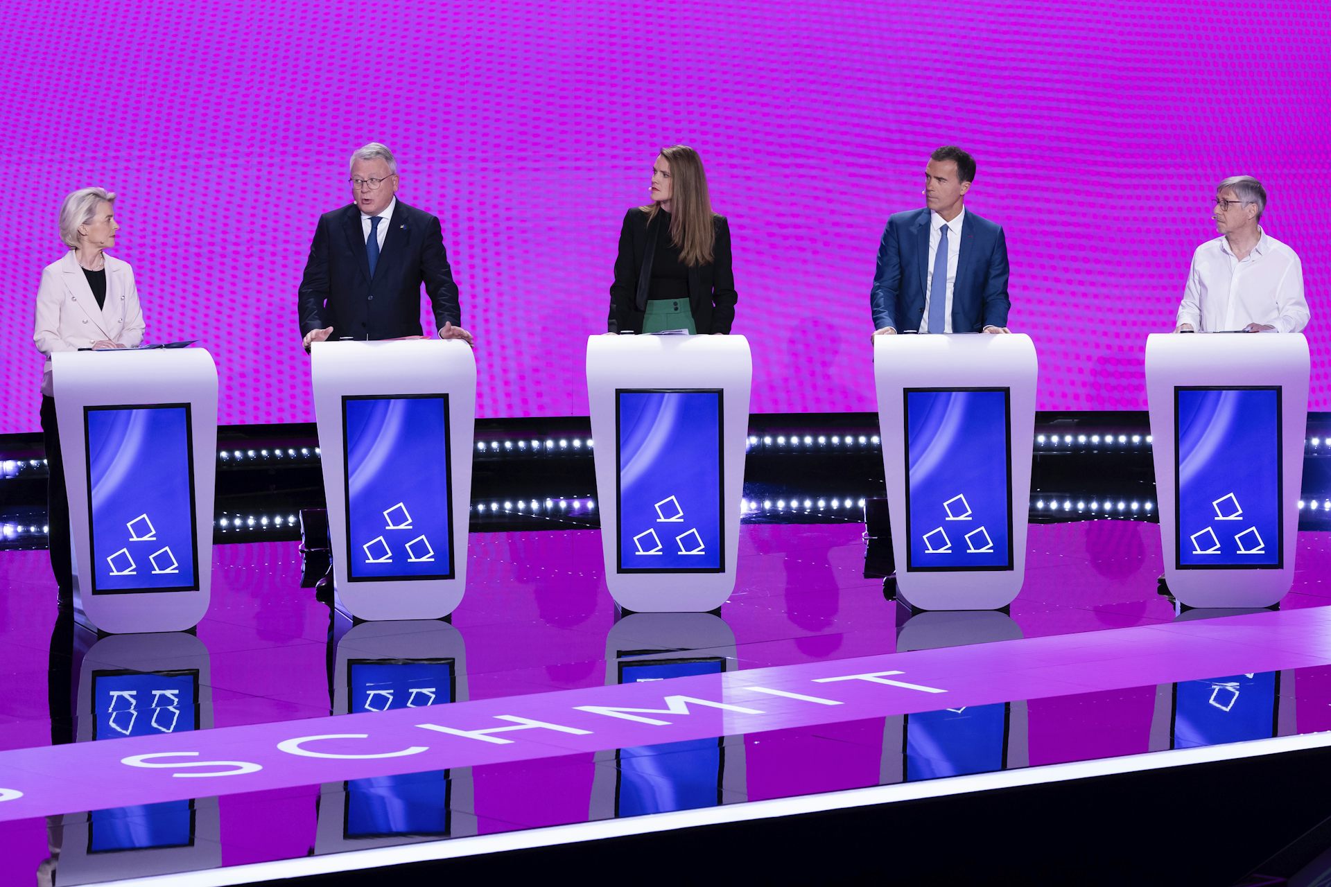 Five politicians stand behind lecterns on a debate stage, with a bright pink backdrop.