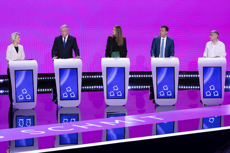 Five politicians stand behind lecterns on a debate stage, with a bright pink backdrop.