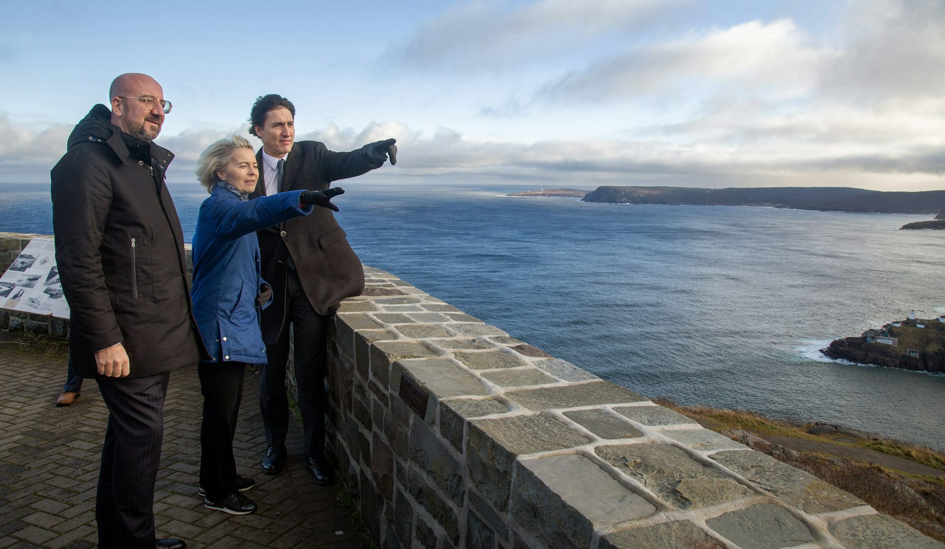 A dark-haired man points to something off camera to a woman and a man. A stone wall is beside them, and the ocean in the background.