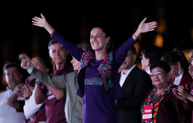 A woman in a purple dress holds her hands aloft in celebration.
