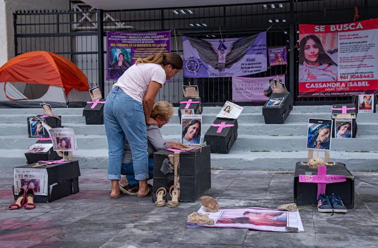 Two women kneel on a pavement surrounded by makeshift graves.