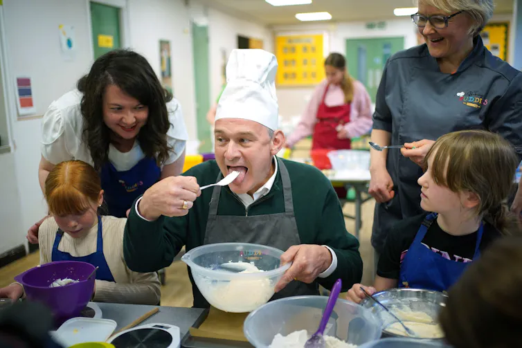Ed Davey wearing a chef's hat and licking a spoon of cake mixture on a visit to a school.