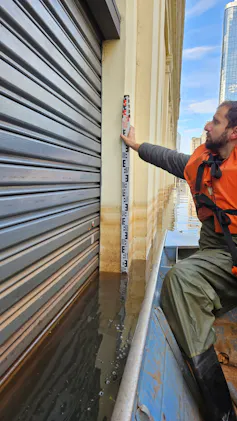 Foto mostra hidrólogo em um barco, com colete laranja, segurando uma régua junto a uma porta de metal. É possível ver a água cobrindo parte da porta e marcas de lama na parede, acima do nível da água