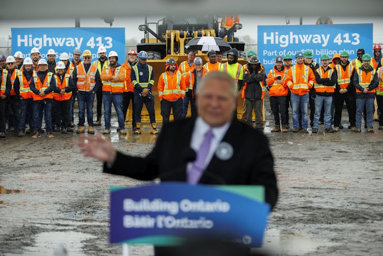 A man in the foreground behind a lectern is blurred with rows of tradespeople behind him and Highway 413 signs in sharp focus.