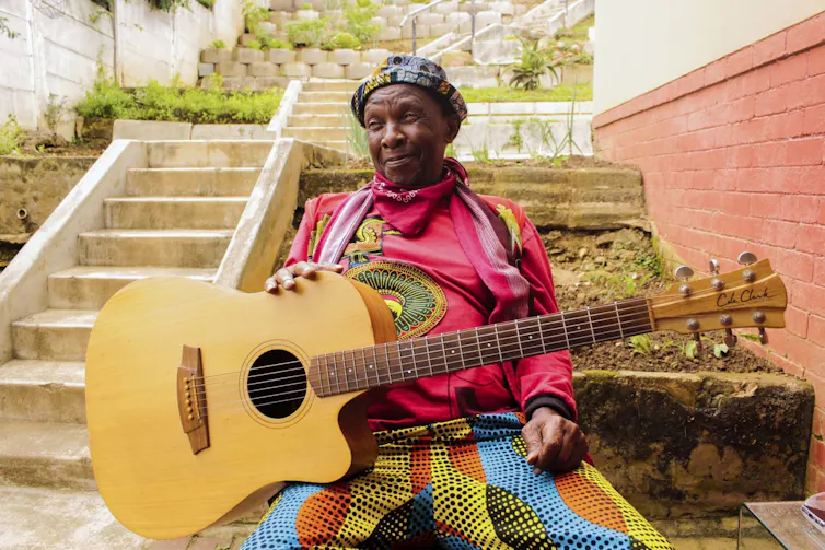 A man in a brightly-coloured jersey sits with a guitar resting on his lap near an outdoor staircase.