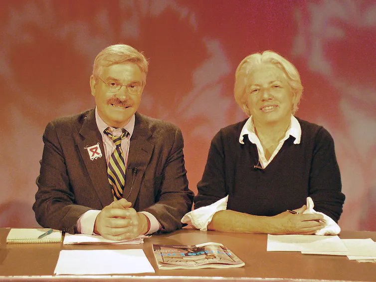 Middle-aged man with mustache and middle-aged woman with short hair sit at a news desk.