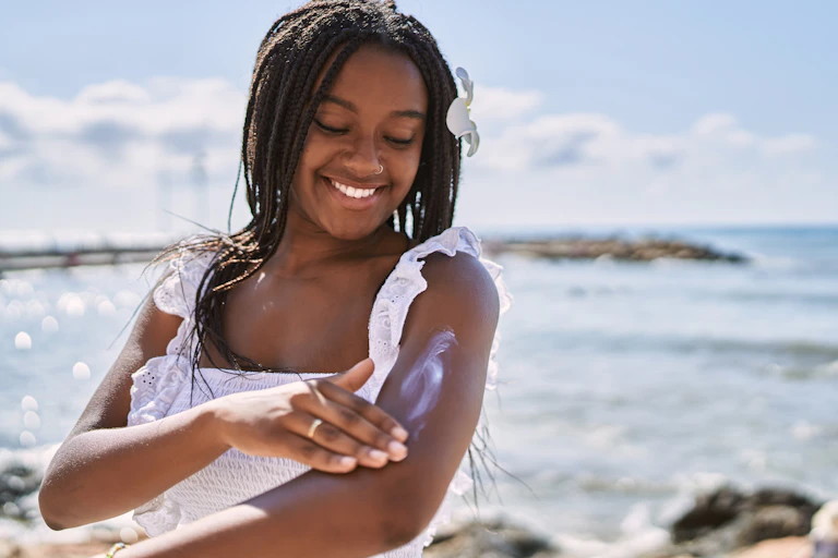 Young pretty black girl with white top and flower in hair applying white sun scream to her arm, sea in background, blue sky