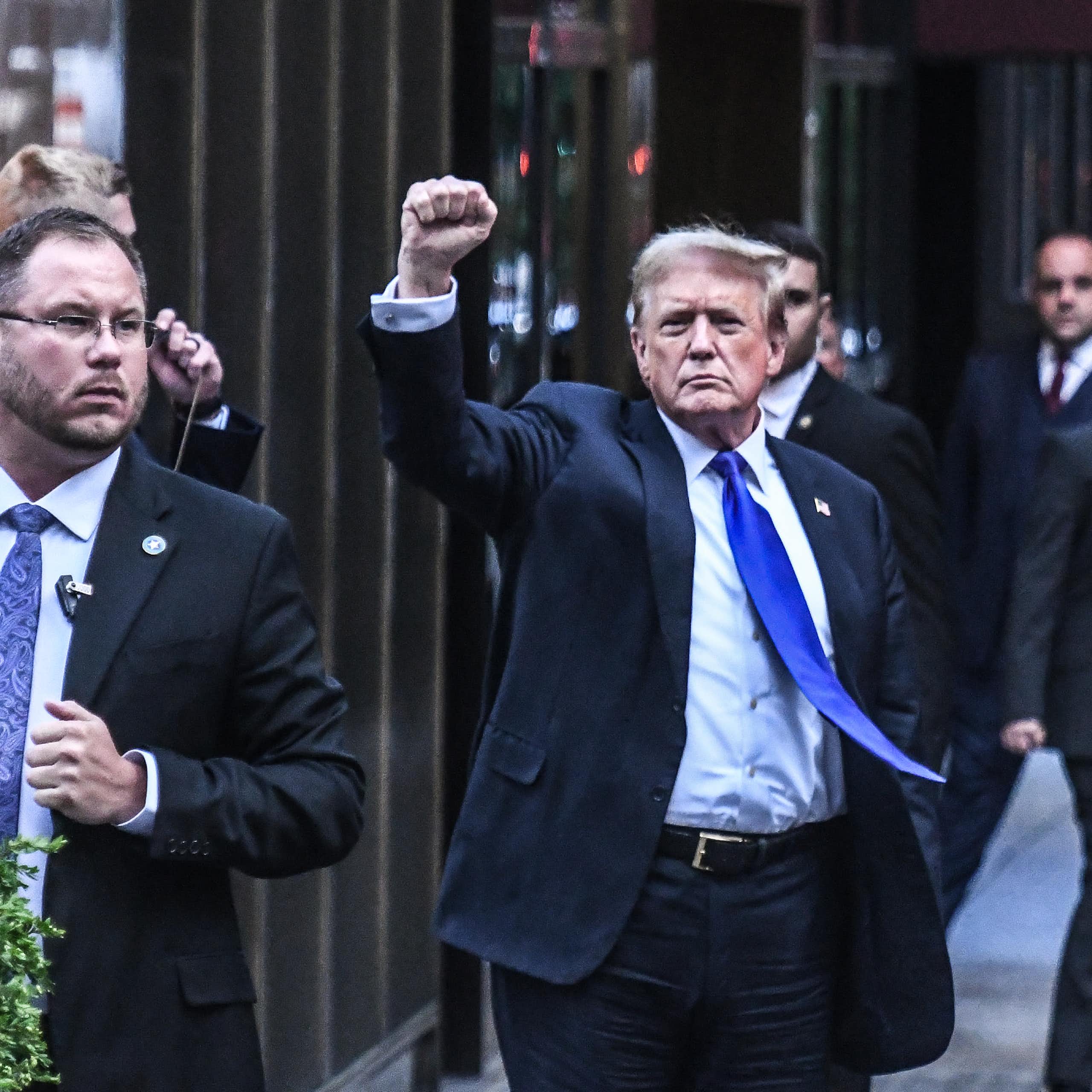 A man in a blue blazer and blue tie raises his fist as he walks on a sidewalk.