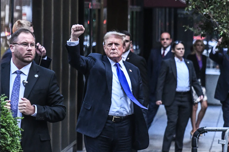 A man in a blue blazer and blue tie raises his fist as he walks on a sidewalk.