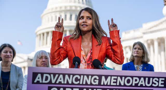 An elegant woman in a dark orange jacket makes air quotes with her fingers against a backdrop of the U.S. Capitol.
