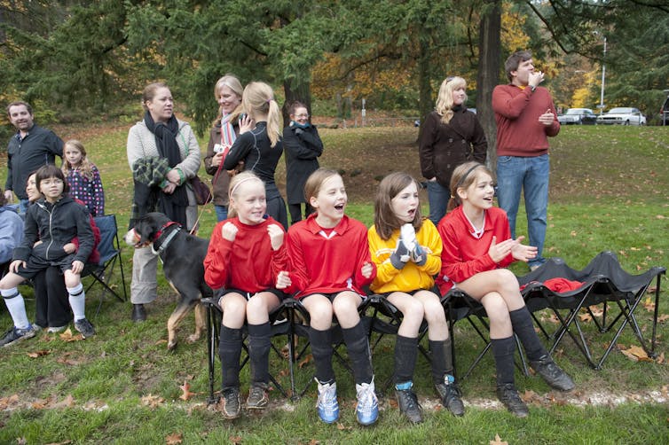 Four uniformed girls sit on the bench while their parents stand around in the background