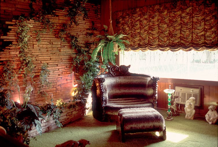 Sala de estar con paredes revestidas de madera, paredes de imitación de piedra, plantas artificiales y alfombras verdes de pelo largo.