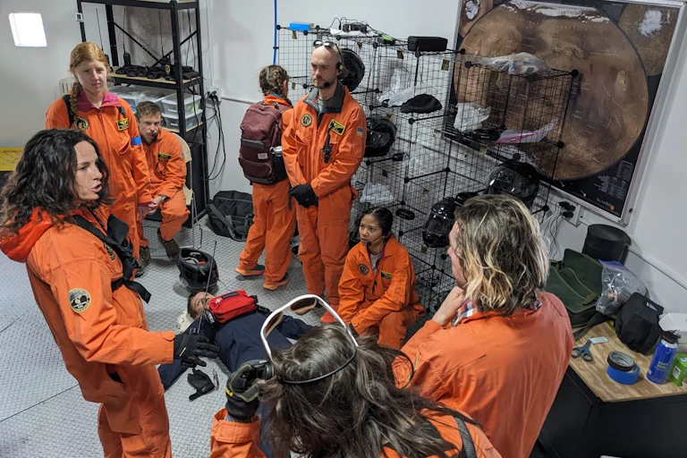 A group of people wearing orange jumpsuits stand and talk while a man in blue jumpsuit lies on the ground.