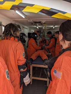 A group of people in orange jumpsuits stand around a table, with a person laying on it.
