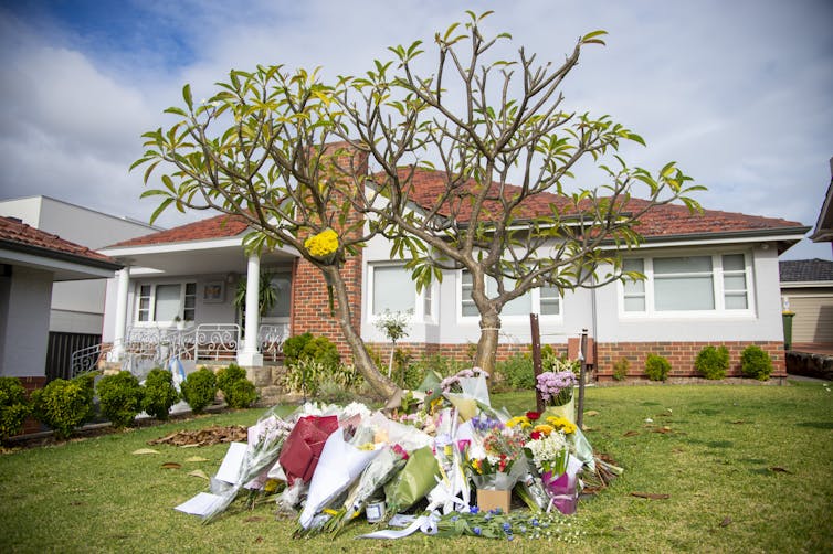 A weatherboard house with dozens of bunches of flowers laid on the front lawn