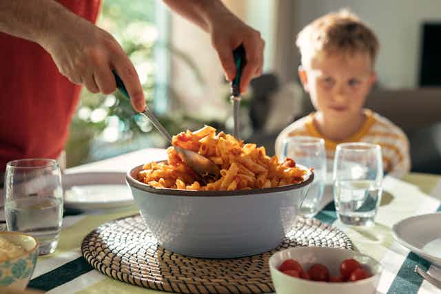 boy in background looks unsure as hands spoon big bowl of pasta at dinner table
