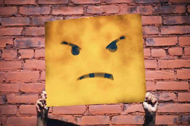 Some hands holding a large yellow sign with an angry face on it