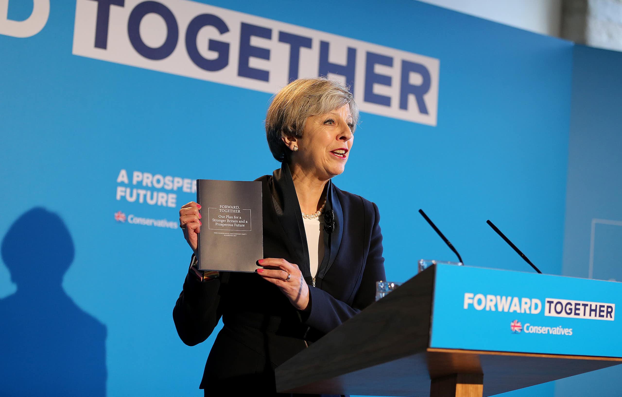 Theresa May, standing at a blue podium reading 'forward together', holds up a copy of the 2017 Conservative manifesto