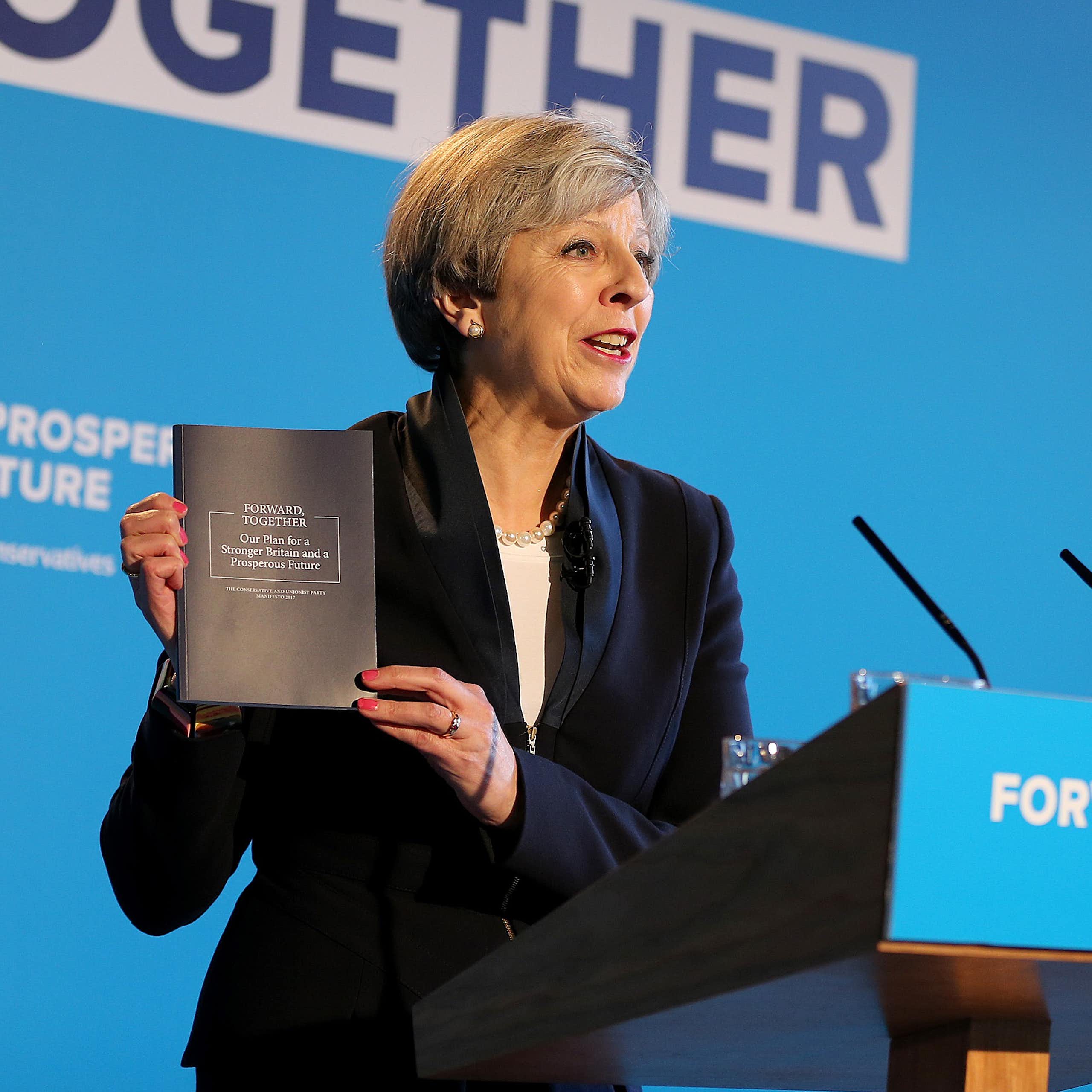 Theresa May, standing at a blue podium reading 'forward together', holds up a copy of the 2017 Conservative manifesto