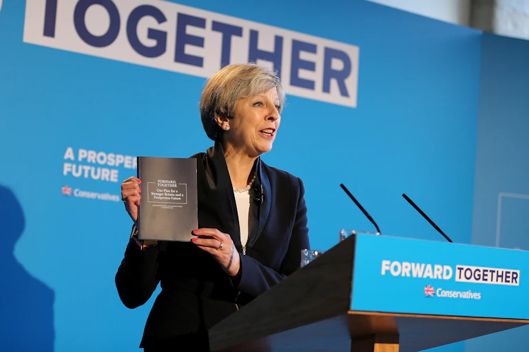 Theresa May, standing at a blue podium reading 'forward together', holds up a copy of the 2017 Conservative manifesto