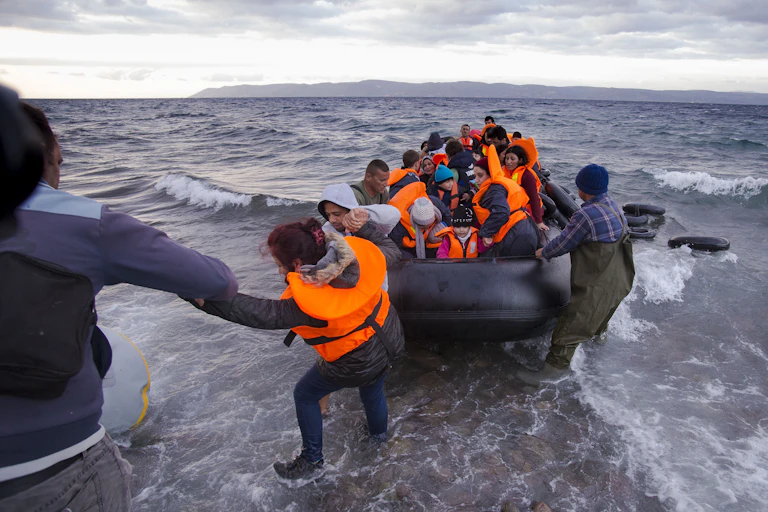 People on shore help pull a rubber dinghy full of people in life jackets from the ocean
