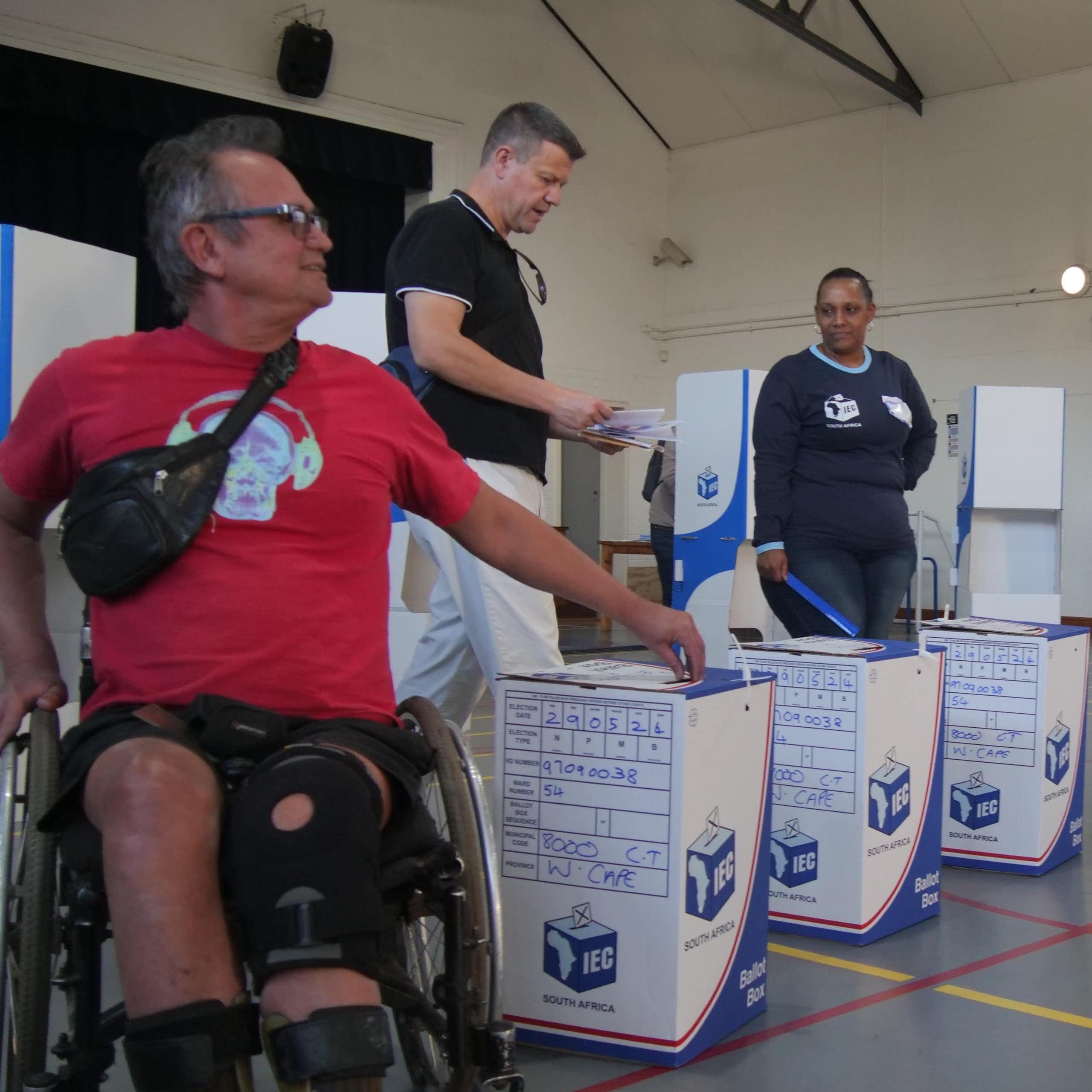 A man in a wheelchair and another standing man in a hall with ballot boxes; an official observes them