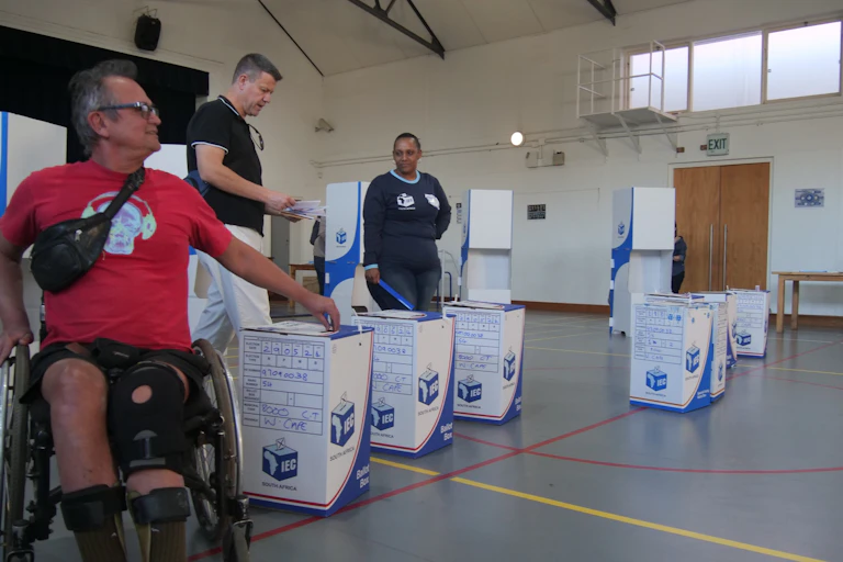 A man in a wheelchair and another standing man in a hall with ballot boxes; an official observes them