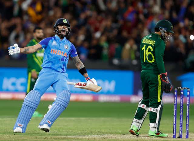Cricketer wearing blue uniform and helmet screams in joy next to a dejected-looking man wearing green.