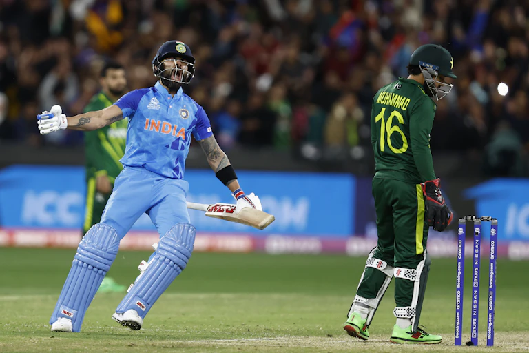 Cricketer wearing blue uniform and helmet screams in joy next to a dejected-looking man wearing green.